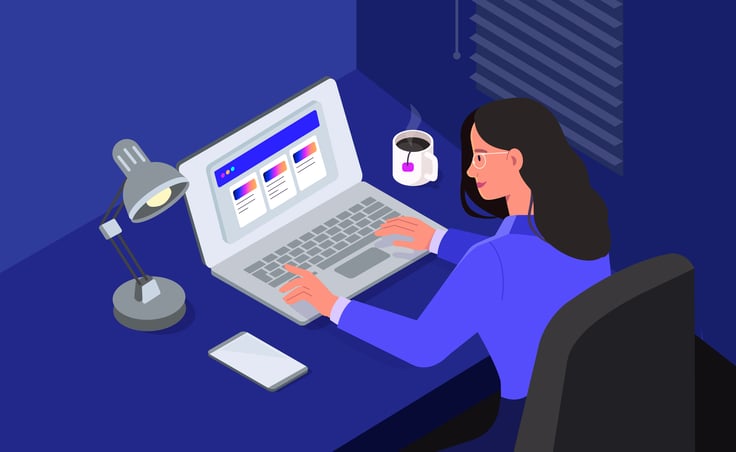 woman wearing glasses working on a laptop at a desk with a lamp, a phone, and a cup of tea